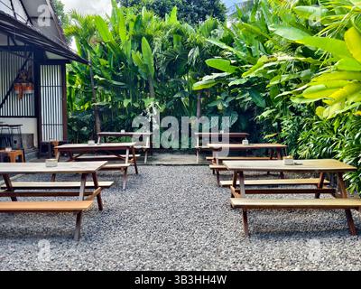 Area salotto all'aperto con diversi tavoli da picnic in legno su un terreno coperto di ghiaia, circondato da lussureggianti piante tropicali Foto Stock