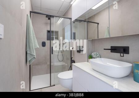 Bagno privato moderno con finiture nere, WC galleggiante, lavandino e cabina doccia in vetro con pareti piastrellate. Foto Stock