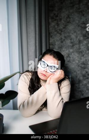 Donna d'affari stanca con occhi dipinti su note adesive affaticate e mal di testa durante il lavoro in ufficio. Sensazione femminile stressata e esausta Foto Stock