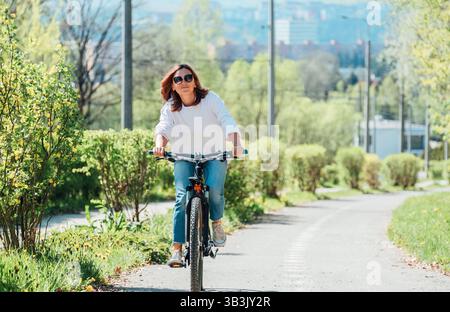 Felice sorridente donna caucasica con eleganti occhiali da sole in bicicletta sulla strada del parco cittadino. Ecologia trasporto sostenibile e Happy People Concept ima Foto Stock