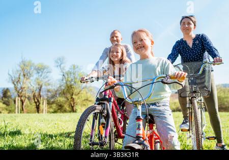 Padre e madre sorridenti con le loro due figlie durante le gite estive in bicicletta all'aperto. Si godono momenti di qualità insieme in un parco cittadino soleggiato. Felice p Foto Stock