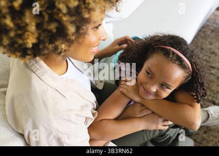 Sorridendo, madre e figlia diverse che si coccolano sul divano nel soggiorno, con cuscino e tappeto Foto Stock
