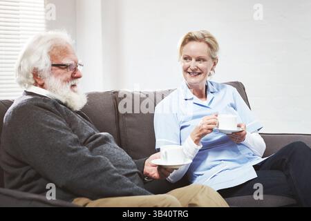 Una coppia matura felice che fa una pausa caffè o tè sul divano di casa. Anziani spensierati e pensionati che si legano, trascorrono una giornata tranquilla insieme, parlano e guardano Foto Stock