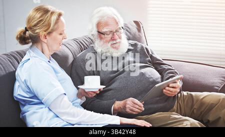 Una coppia matura che guarda un tablet digitale sul divano, si siede, parla e si rilassa a casa. Vecchio uomo anziano che legge online il suo partner, moglie o infermiera Foto Stock