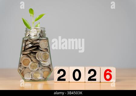 Business e New Year Concept 2026. Parola 2026 scritta su cubo bianco o blocco con monete in bottiglia. Concetto di investimento e risparmio. Le piante giovani crescono Foto Stock