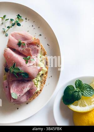 Toast all'avocado con prosciutto, erbe aromatiche e limone su sfondo bianco. Cibo sano. Direttamente sopra, copia spazio. Foto Stock