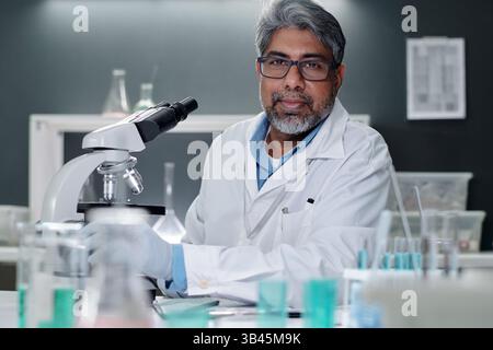 Ritratto di scienziato anziano che indossa camice da laboratorio e occhiali che lavorano con il microscopio in un moderno laboratorio con varie provette e attrezzature che lo circondano Foto Stock