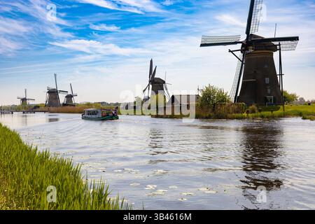 Vista panoramica di Kinderdijk, Paesi Bassi, con storici mulini a vento lungo un canale, con una barca con persone sull'acqua e sponde verdi sotto un BL Foto Stock