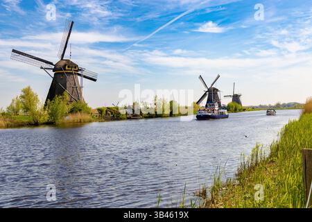 Vista panoramica di Kinderdijk, Paesi Bassi, che mostra numerosi mulini a vento storici lungo un ampio corso d'acqua con una barca, con sponde verdi e un cielo blu Foto Stock