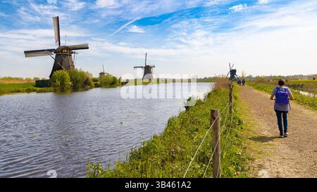 Vista panoramica di Kinderdijk, Paesi Bassi, con storici mulini a vento lungo un sentiero vicino all'acqua, con una persona che cammina, campi verdi e un cielo blu Foto Stock