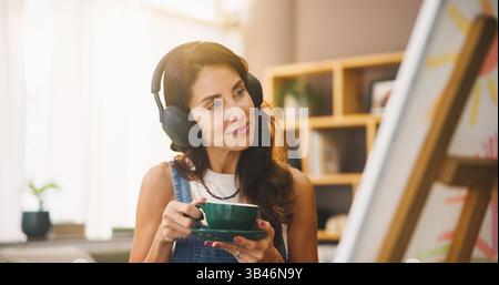 Donna, artista e caffè con cuffie per dipingere, ascoltare musica e progetti artistici. Persona, bevanda e audio in streaming con canvas per Foto Stock