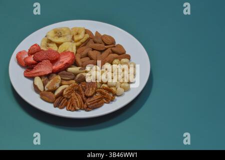 Una varietà di frutta secca e frutta secca su un piatto su sfondo blu. Nocciole, noci pecan, noci del brasile, mandorle, fragole secche e banane. Il concetto Foto Stock