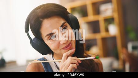 Pensiero, donna e pittrice con cuffie a casa per lo streaming di musica, podcast audio e idee artistiche. Sorridi, persona e pensieri con pennello per Foto Stock