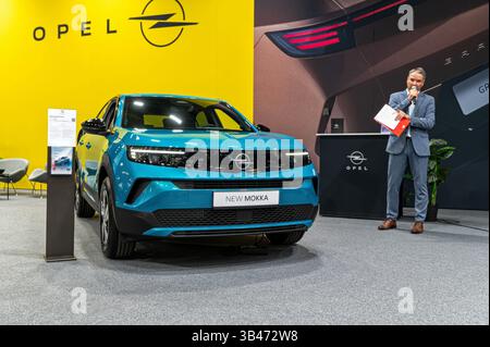 Riga, Lettonia - 25 aprile 2025: Un presentatore parla della nuova Opel Mokka in un salone di auto con sfondo giallo brillante. Foto Stock