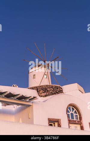 Santorini, Grecia - 15 ottobre 2024: Classico mulino a vento di Santorini al tramonto con cielo blu e tradizionale architettura bianca. Foto Stock