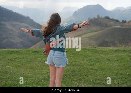 Vista posteriore di una giovane donna in abiti estivi che abbraccia il paesaggio montano con le braccia allungate. Il vento nuvoloso crea una scenografia all'aperto spettacolare. Foto Stock