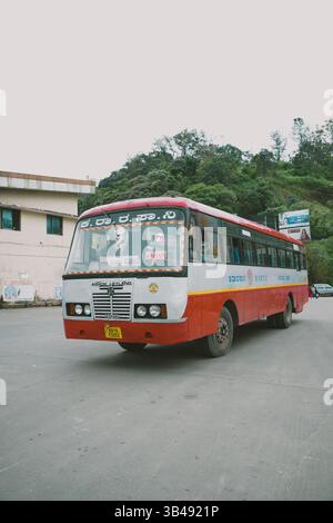 Un autobus rosso e bianco KSRTC Ashok Leyland è parcheggiato su una strada in una zona collinare, con vegetazione ed edifici sullo sfondo sotto un cielo coperto. Foto Stock