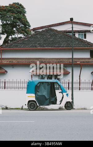 Un risciò parcheggiato in una tranquilla strada indiana accanto ad una casa con tetto piastrellato, con alberi in fiore che creano una vibrante atmosfera urbana quotidiana. Foto Stock