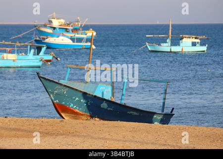 Barca da pesca indossata sulla costa sabbiosa di Safaga, Egitto, con navi ormeggiate nel Mar Rosso sotto la luce del pomeriggio. Foto Stock