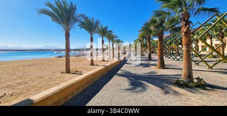 Passeggiata panoramica pedonale lungo la spiaggia sabbiosa di Sahl Hasheesh, Egitto, con palme, ombrelloni e vista sulla costa del Mar Rosso. Foto Stock