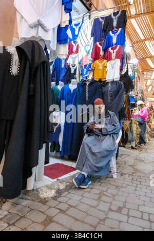 30 DEC 2024, Marrakech, Marocco - Un commerciante di mercato in un piccolo bazar di souk in attesa di clienti. Vendere vestiti Foto Stock