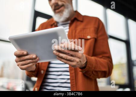 Un uomo anziano con la barba interagisce con un tablet mentre si trova in un ufficio moderno. Foto Stock