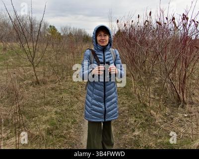 La donna rimane immobile, scansionando attentamente gli uccelli nella foresta e nel prato sparso di inizio stagione. Foto Stock