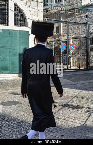 Ebreo ultra-ortodosso con abiti tradizionali che camminano in una città vecchia di Gerusalemme. Vista posteriore dell'uomo assidico che indossa cappelli in pelliccia shtreimel che cammina su una strada Foto Stock
