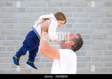 Un nonno gioioso solleva il suo bambino ridendo in alto davanti a un muro di mattoni chiari. Foto Stock