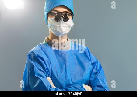 Ritratto di chirurgo femminile in scrub blu e maschera in piedi con le braccia incrociate in sala operatoria. Foto Stock