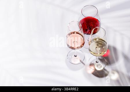 Tre bicchieri da vino pieni di vino rosso, bianco e rosa, in piedi graziosamente su uno sfondo luminoso e minimalista. Una vivace esposizione di varietà di vini Foto Stock