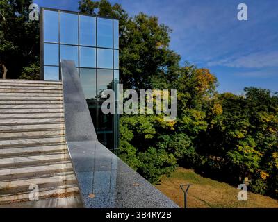 Ampia scala in marmo e ringhiera in pietra lucida che conduce ad un alto e moderno edificio in vetro, circondato da lussureggianti alberi autunnali verdi e gialli sotto un cle Foto Stock