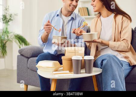 Coppia sorridente seduta sul divano divertendosi a chiacchierare e gustando il cibo consegnato. Foto Stock