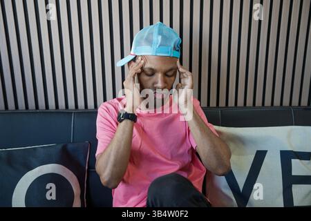 Giovane che tocca la testa e soffre di un forte mal di testa mentre si siede su un divano Foto Stock