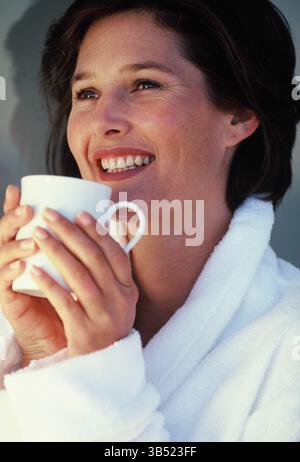 Primo piano di una giovane donna all'interno in accappatoio, con una tazza in mano. Foto Stock