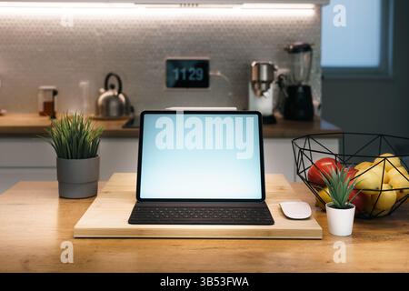 Tavoletta digitale con mockup a schermo vuoto, piano cucina in legno, casa accogliente e moderna. Frutta fresca, piante decorative, elettrodomestici da cucina e simboli Foto Stock