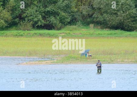 Una tranquilla scena sul fiume Loira in Francia, che cattura un uomo lontano in piedi in acqua mentre pesca. Sua moglie si rilassa sulla riva del fiume. Foto Stock