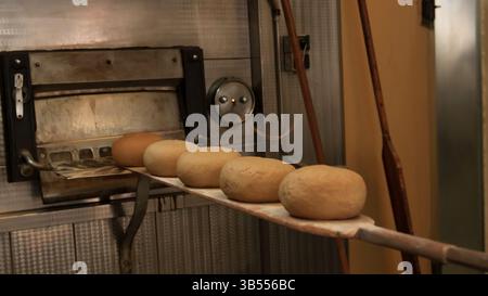 Primo piano dell'impasto di pane crudo posto su una paletta di legno, pronto per essere cotto in un forno industriale. La scena cattura il processo di cottura tradizionale in un Foto Stock