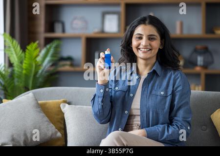 Ritratto di una giovane donna musulmana sorridente seduta sul divano a casa in una camicia, che tiene in mano un inalatore per respirare, e che mostra una bottiglia di medicina alla macchina fotografica. Foto Stock