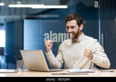 Giovane uomo felice di successo e risultato che mostra un gesto positivo con le mani, seduto in ufficio alla scrivania, guardando lo schermo del notebook. Foto Stock