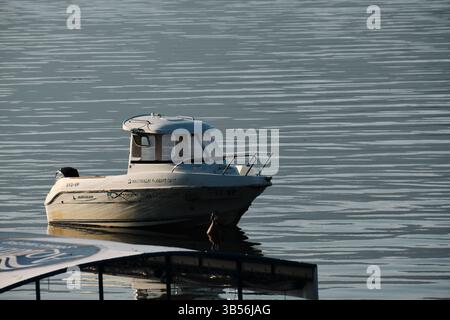 Primo piano di una piccola barca a motore bianca che galleggia sul lago Skadar, in Montenegro, con calme acque ondulate e luce del mattino presto che si riflette sulla superficie Foto Stock