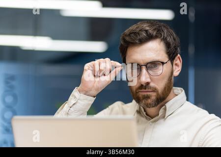 Foto ravvicinata di un giovane uomo d'affari sorpreso seduto in ufficio alla scrivania e guardando con preoccupazione lo schermo del notebook, alzando gli occhiali sul viso. Foto Stock