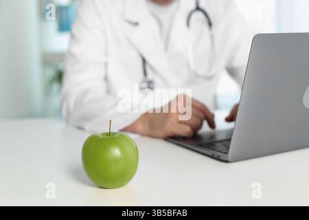 Nutrizionista che lavora con il computer portatile a tavola in clinica, si concentra sulla mela verde Foto Stock