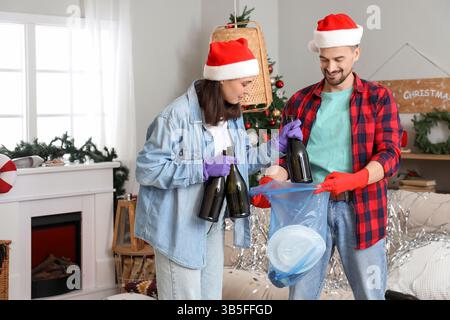 Una giovane coppia di cappelli di Babbo Natale che getta bottiglie in un sacco della spazzatura a casa dopo la festa di Capodanno Foto Stock