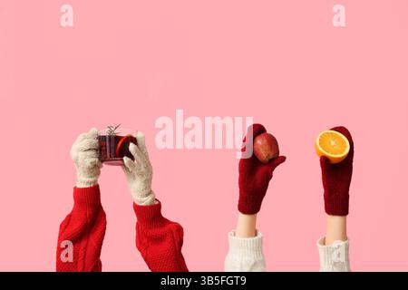 Mani femminili in guanti che contengono melograno, arancia e tazza di gustoso VIN brulé su sfondo rosa Foto Stock