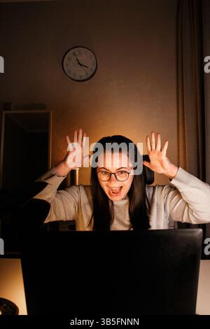 Donna millenaria che celebra la vittoria in casa, donna d'affari che lavora con un notebook tenendo le mani in alto, soddisfatta ha ottenuto ottimi risultati Foto Stock