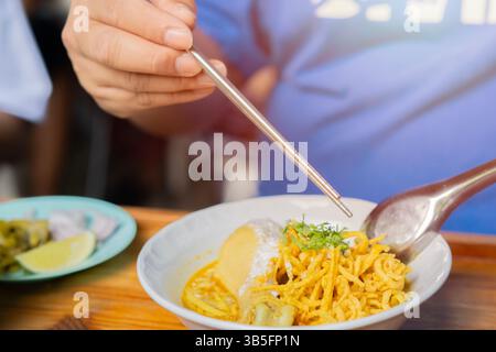 Attenzione selettiva dell'uomo che tiene le bacchette con Khao Soi Kai con scivolo di lime, Khao Soi Kai o tagliatelle tailandesi al curry con tagliatelle all'uovo e drumsti di pollo Foto Stock