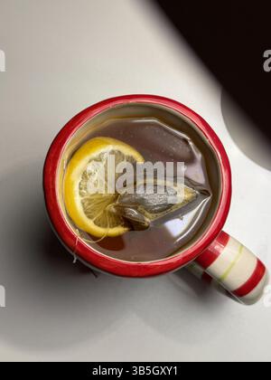 Questa immagine mostra una vista dall'alto verso il basso di una tazza a strisce rosse e bianche riempita di tè caldo. Una bustina di tè e una fetta di limone fluttuano all'interno. La tazza è al posto Foto Stock