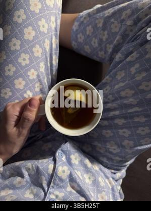 L'immagine mostra una persona in pigiama blu con un motivo floreale bianco, seduta su una superficie scura e con una tazza di tè bianco con una fetta di limone e. Foto Stock