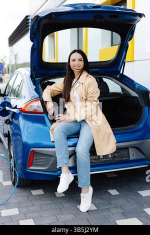 Giovane donna seduta nel bagagliaio di un'auto, in attesa di ricaricarsi e bere un caffè. Foto Stock